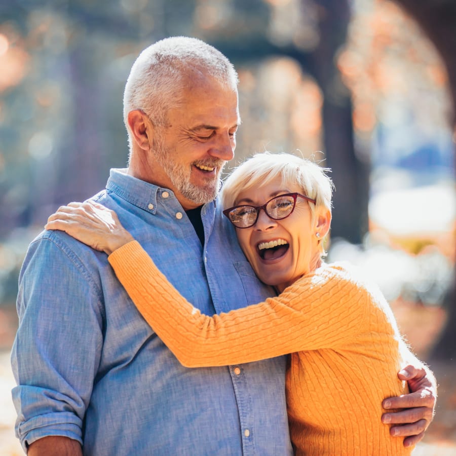 An older couple are laughing while hugging one another on a wooded outdoor trail.