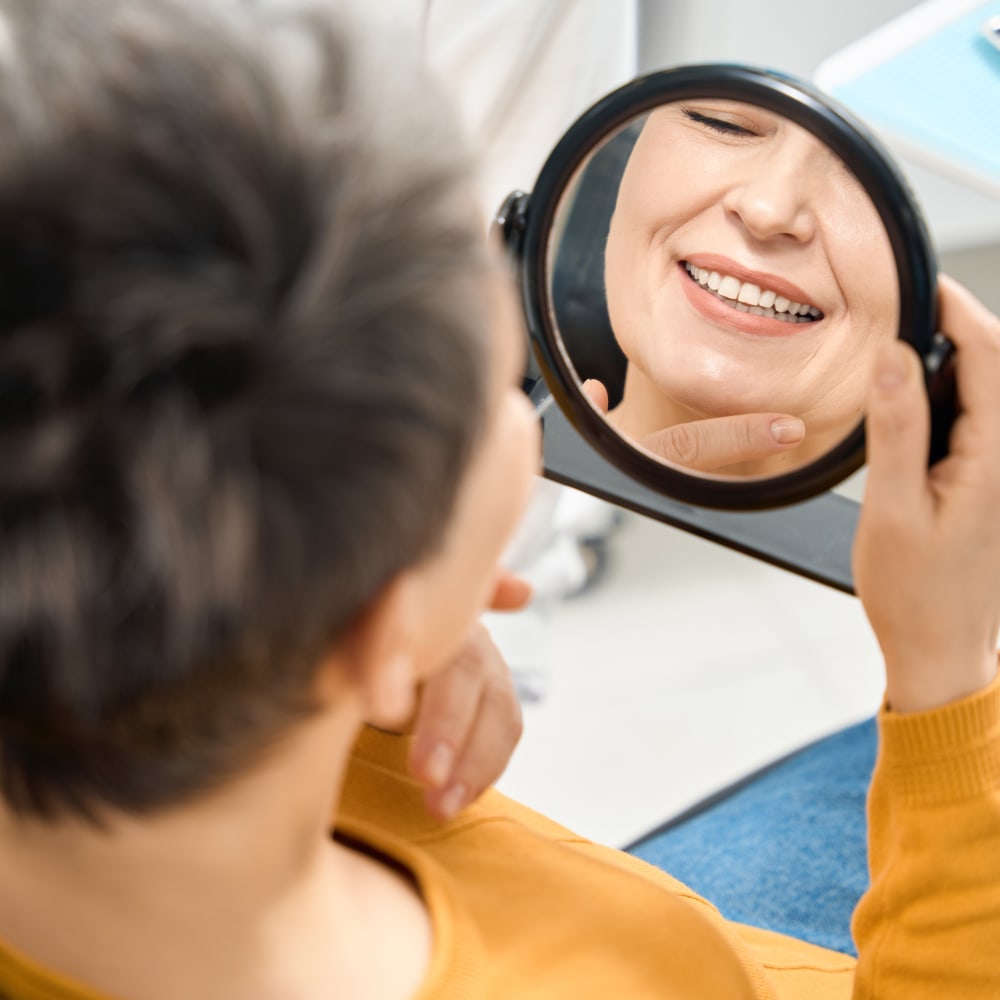 An older lady admires her new smile in a mirror while sitting in a dental chair.