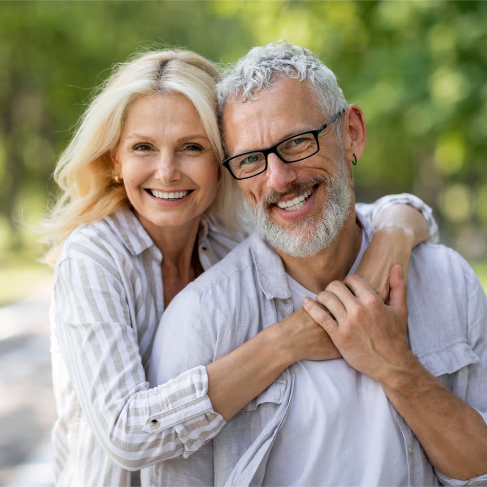 An older woman has her arms around an older man. They are both facing forward and smiling for the camera.