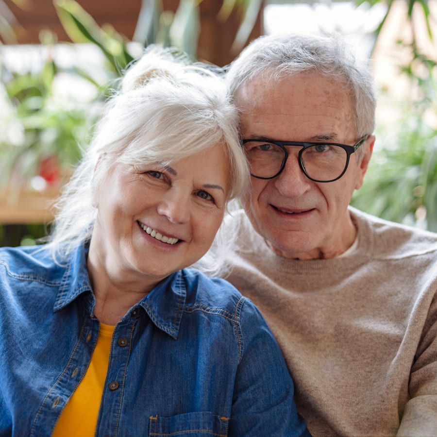 An older couple in a room with lots of plants smile at the camera.