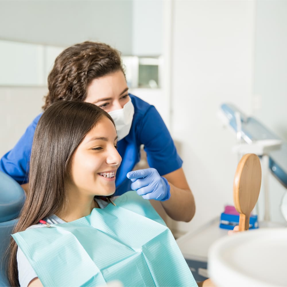 A teen girl is smiling, looking at her braces in a handheld mirror. Her dental professional stands behind her and gestures to her teeth.