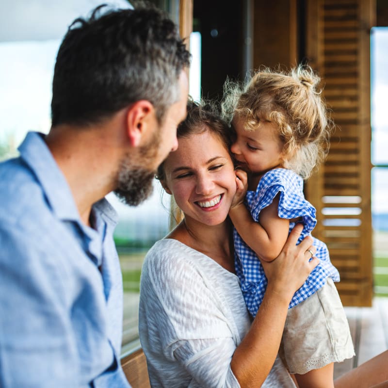 A man looks on as a woman receives a kiss on the cheek from a toddler she is holding.