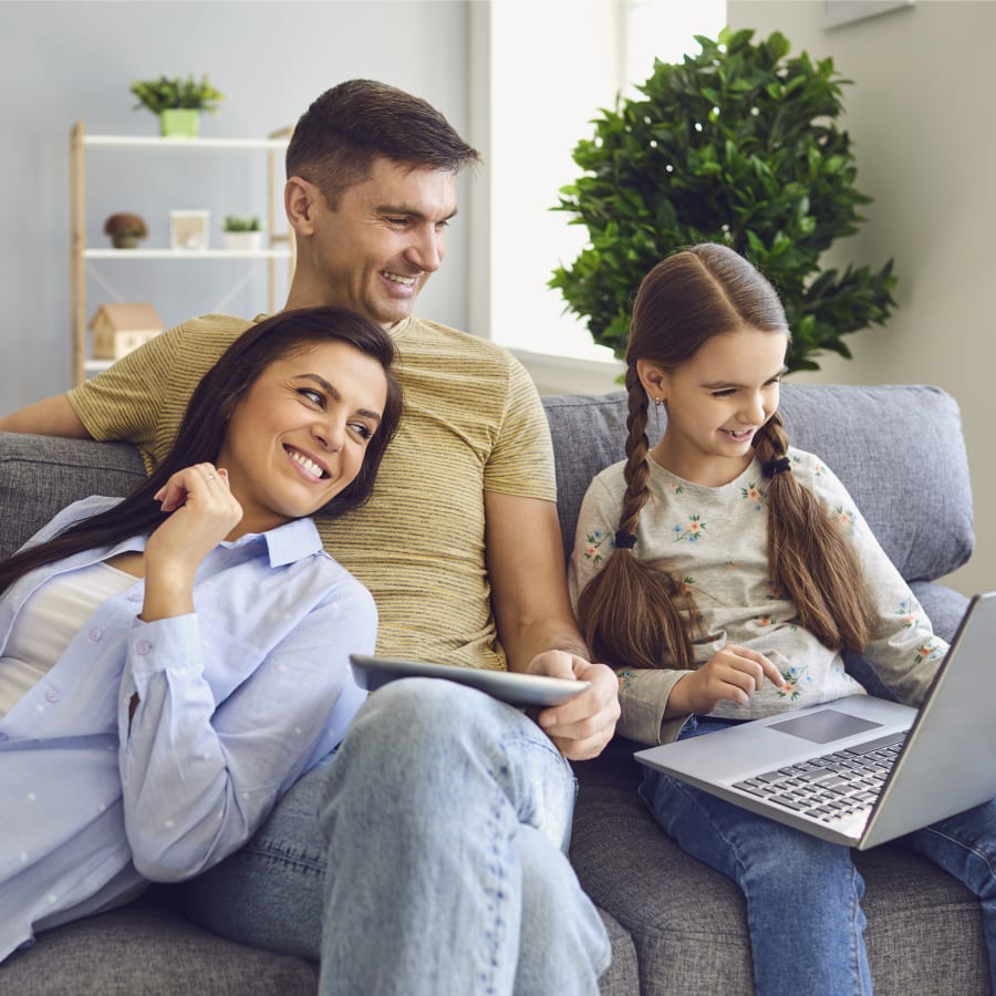 A family of three are sitting on the couch together. The young girl is holding a laptop, and the older man and woman are looking at it with her.