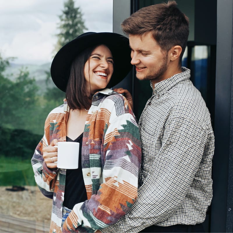 A young couple are standing on their porch smiling at one another while drinking coffee.